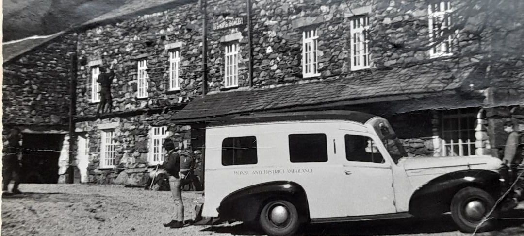 A black and white image from the 1960s of an old ambulance vehicule in front of a slate house.