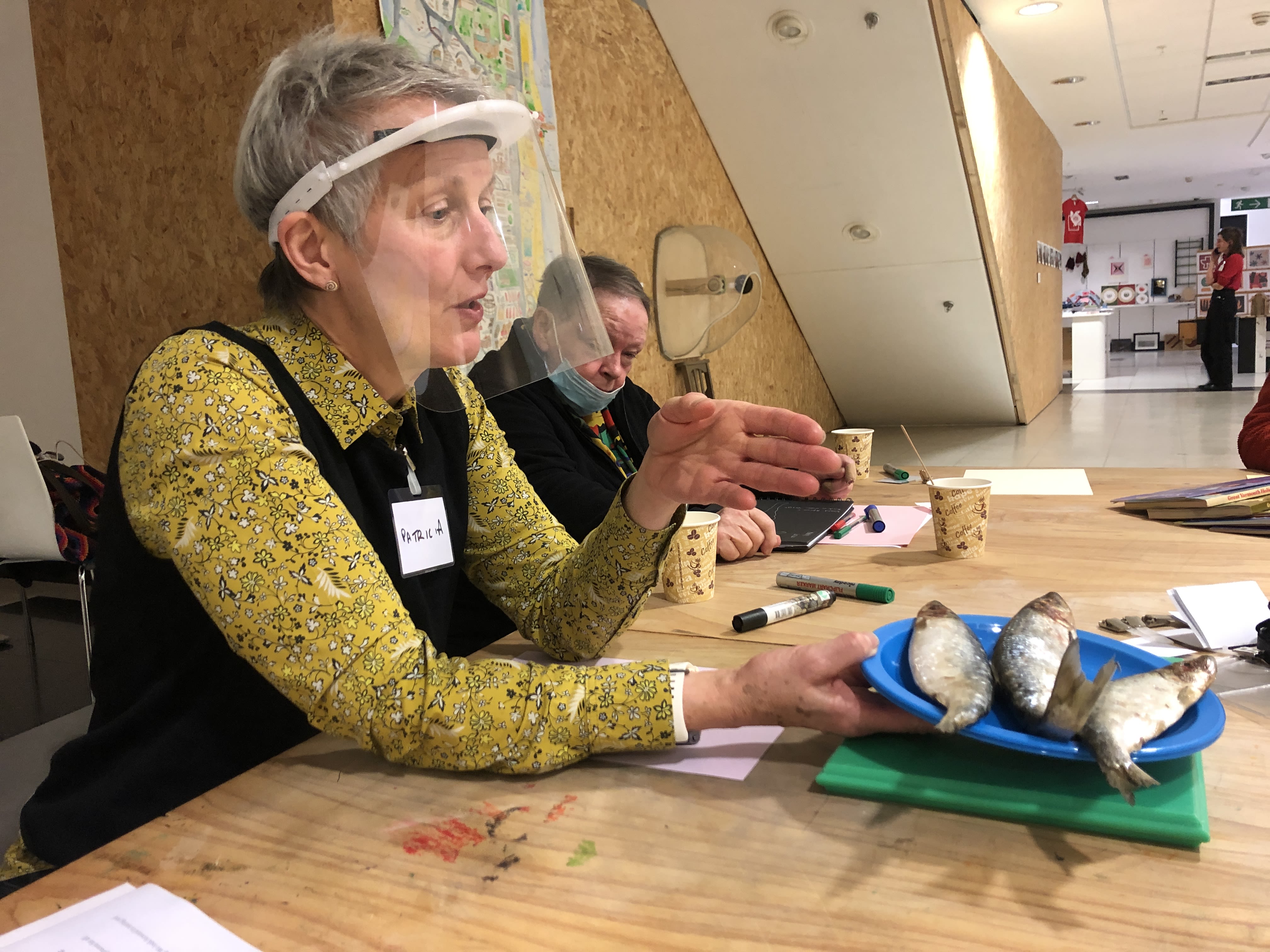 A lady sits at a table holding a plate of herring. She is taking part in a collage workshop.
