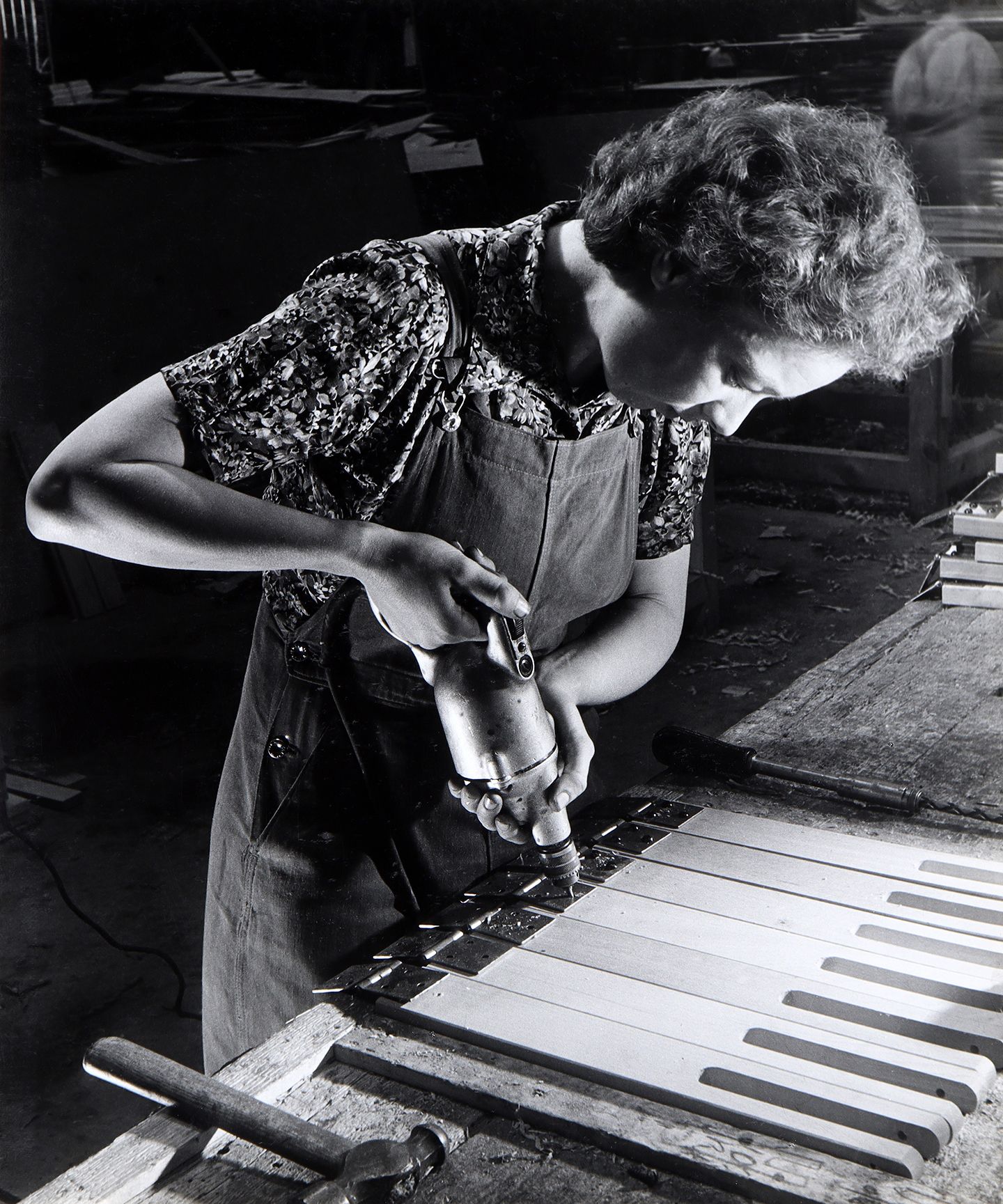 A black and white photo of a woman drilling plot holes in a wood-working factory. She is holding the drill in her right hand.