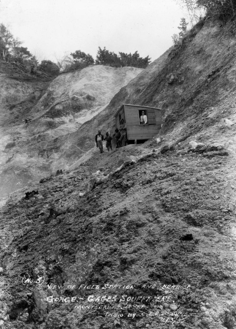 A black and white photo shows a simple hut on a mountain with a man inside and several people outside