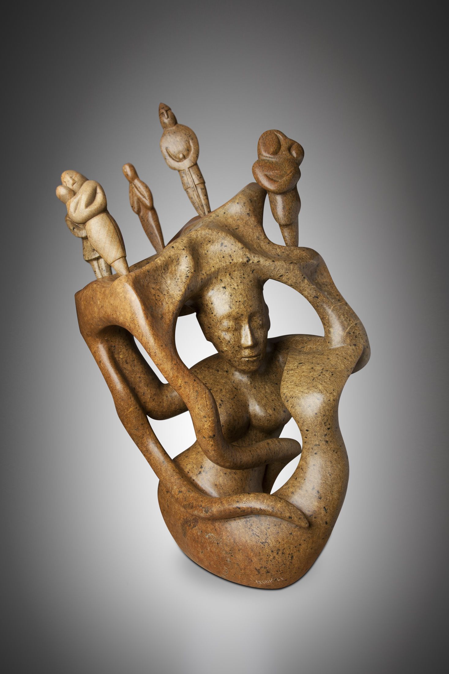 A photo of a soapstone sculpture depicting an abstract human form resembling a mermaid. The human form is holding aloft a platform on which five human forms are stood.