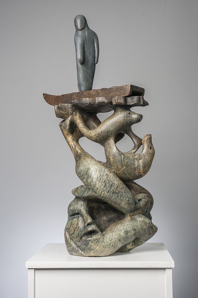 A photo of a soapstone sculpture depicting an abstract human form stood on top of a sled. The sled is mounted on a pile of marine mammals and a human head with an open mouth.