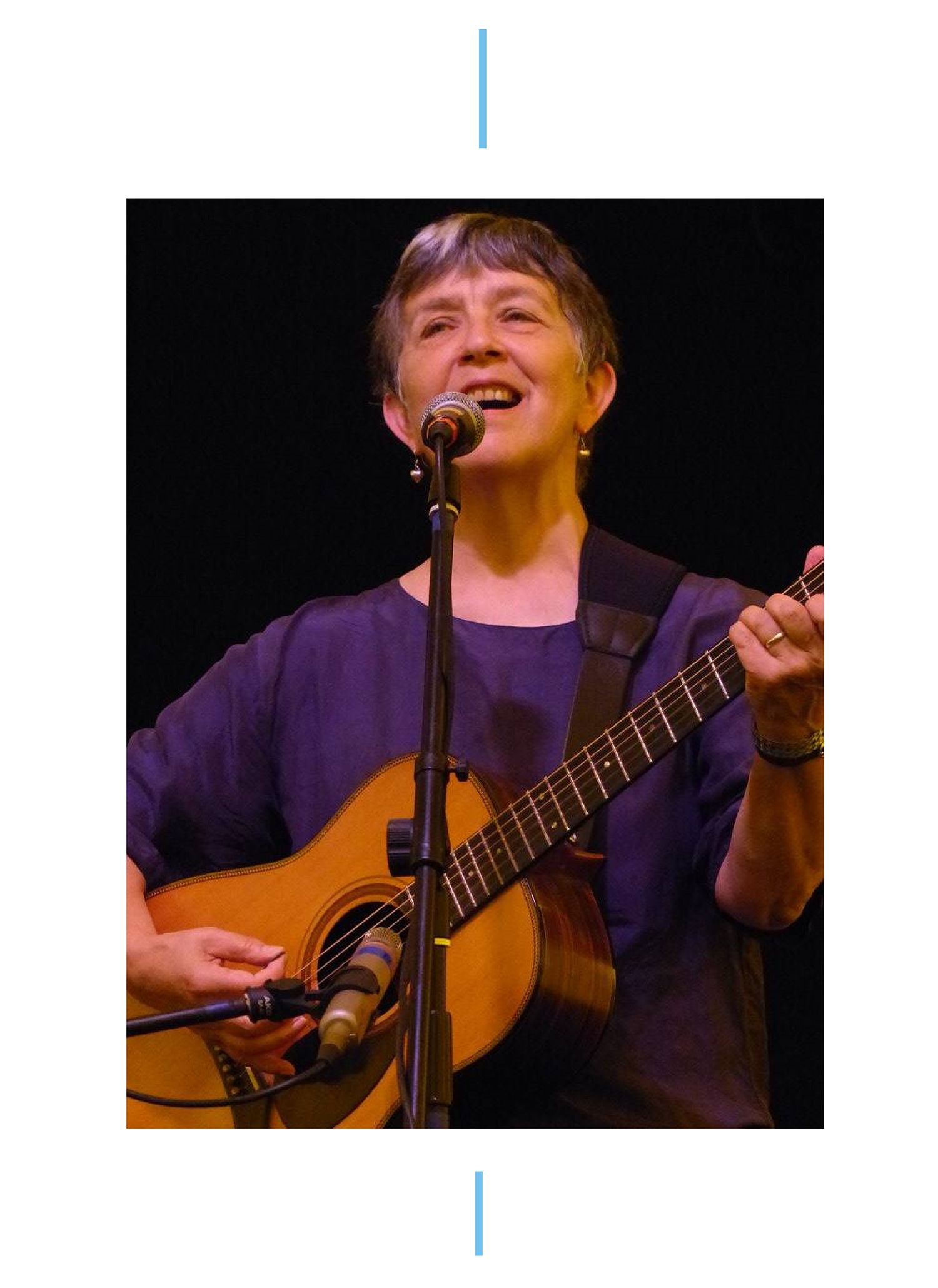 A photo of a female musician in a purple t-shirt singing into a microphone whilst playing an acoustic guitar.