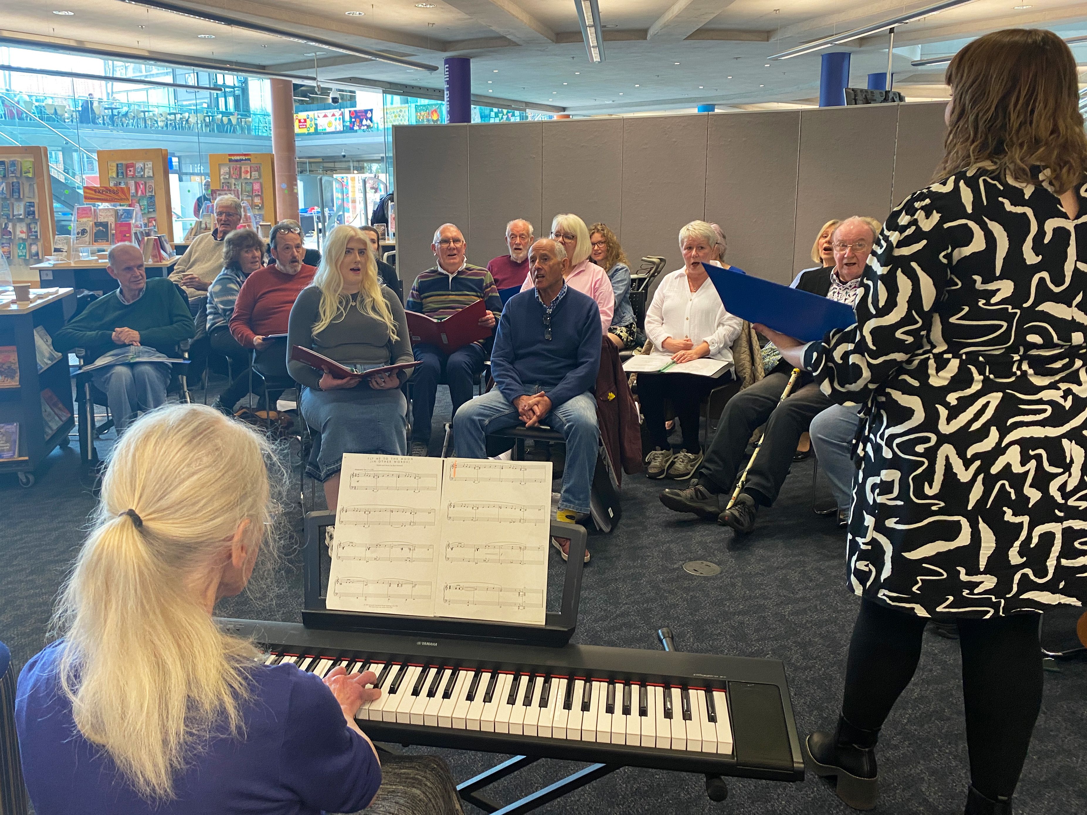 Choir attendees sitting down and singing at the Aphasia Choir, while a person plays music on a keyboard