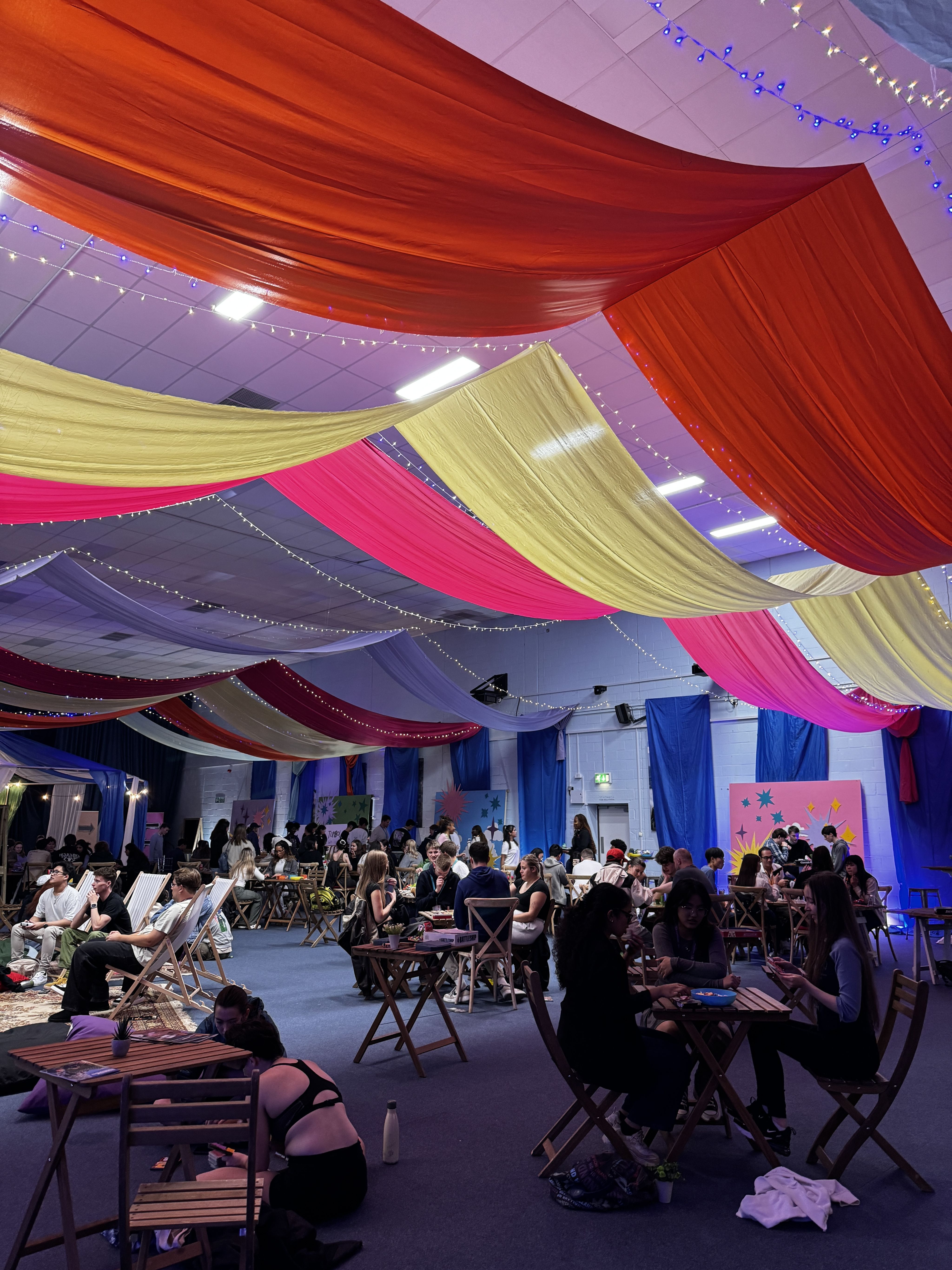 A room with colourful banners hanging from the ceiling and lots of students sitting in chairs
