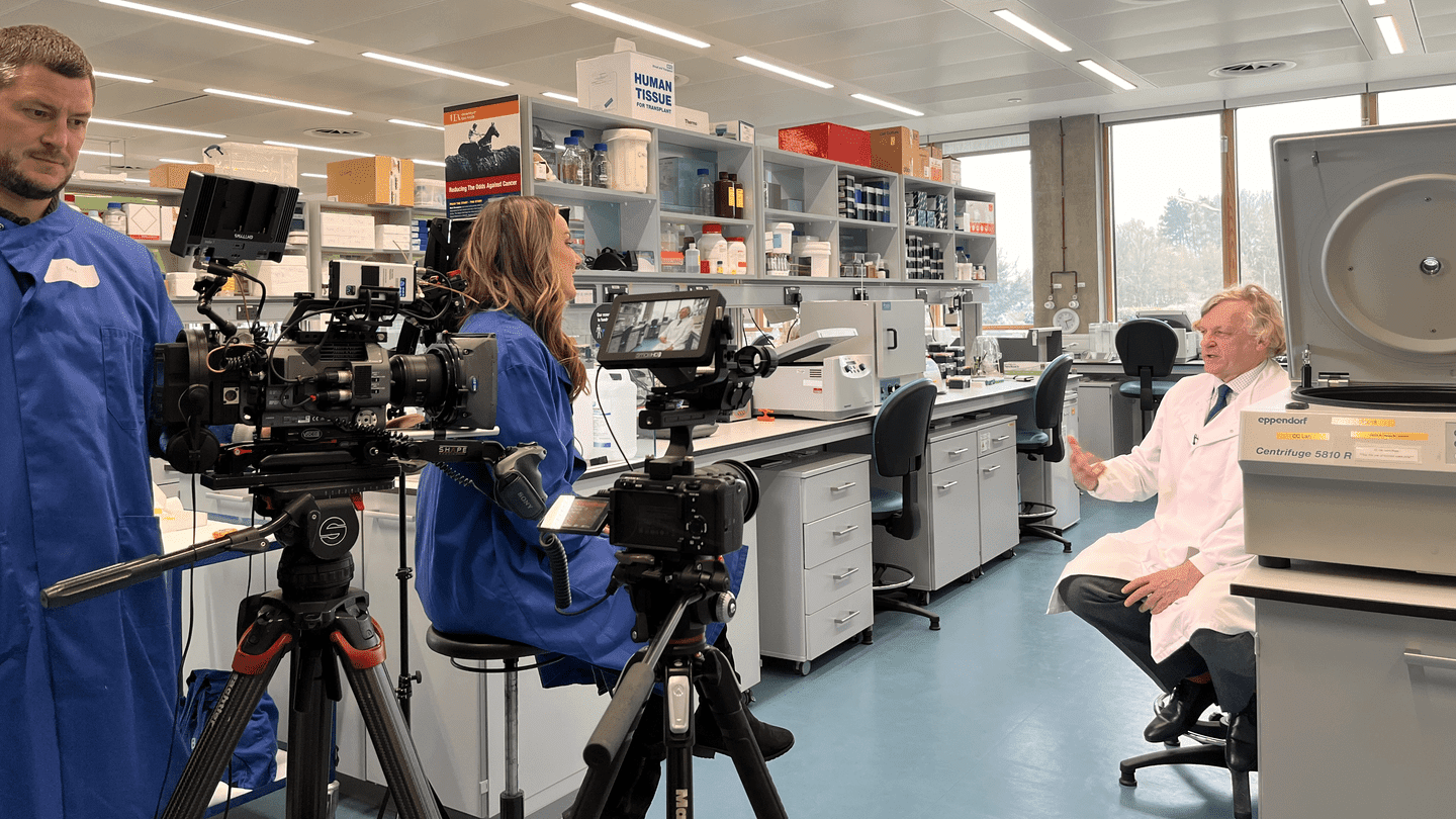 Colin Cooper seated on a chair and wearing a white lab coat, with a camera pointing at him