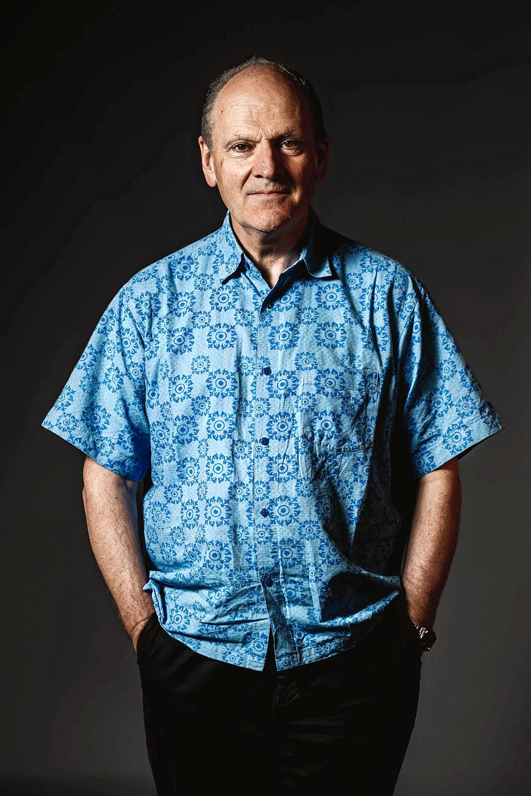Full body image of Prof Robert Nicholls, standing, looking towards the camera with hands in his pockets against a plain black background. He is an older, white gentleman, wearing a patterned blue shirt.