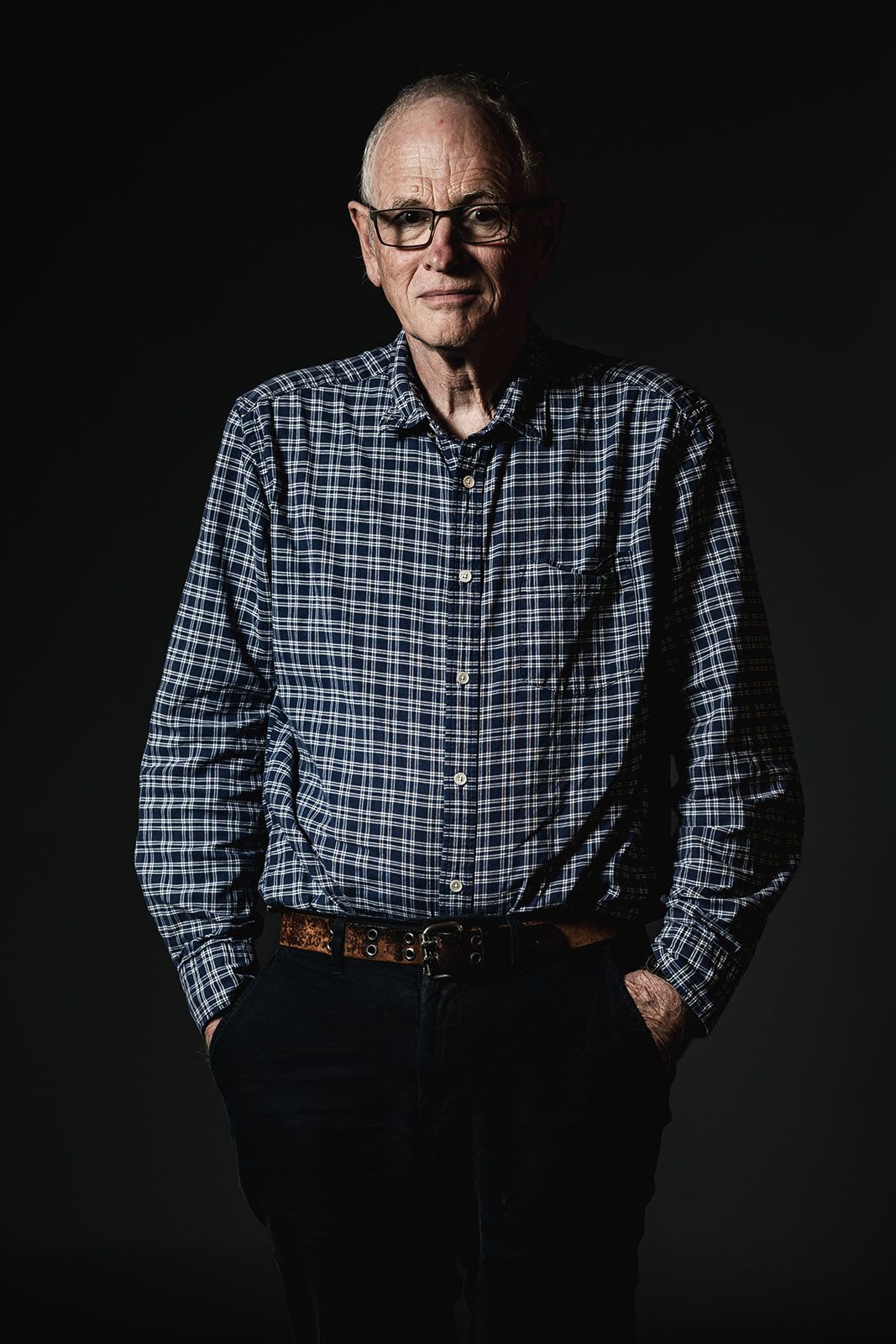 Full body shot of Prof Philip Jones standing against a black background, hands in pockets. Phil is wearing black trousers and a checked shirt. Phil has a serious expression.