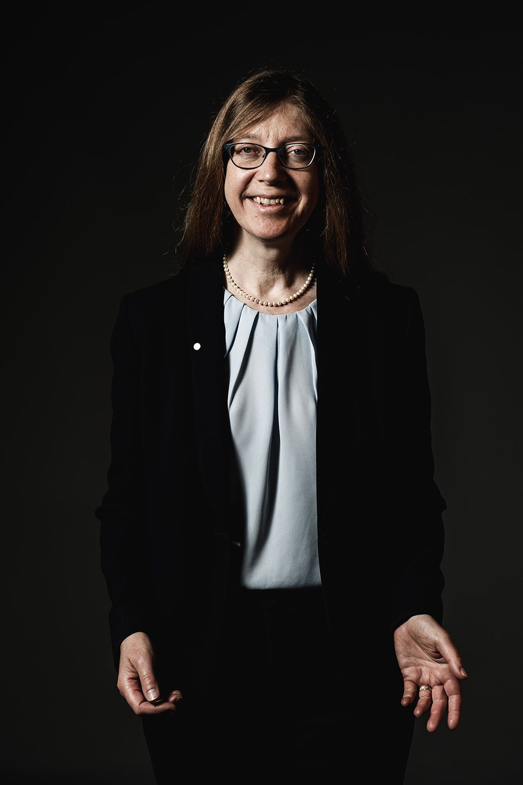 Prof Rachel Warren stands against a black background. She is smiling with an open hand gesture. She is wearing a black suit with a light blue top under the jacket