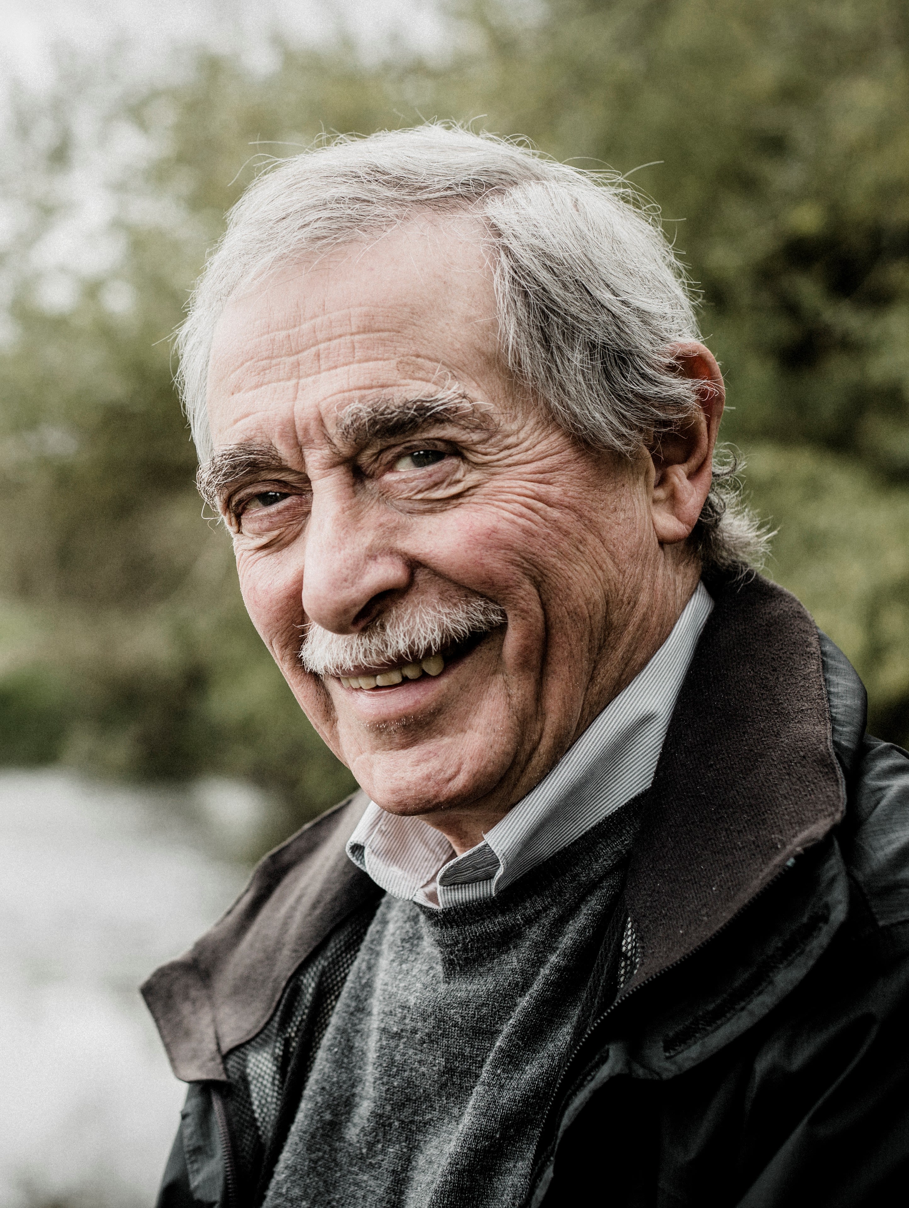 Headshot of Prof Peter Liss, in half profile, smailing at the camera. He is standing ourside with a blurry background of trees and a river behind him. He's an elderly man with grey hair and a short moustache. he's wearing a dark green waxed jacket, green jumper and white shirt. ,